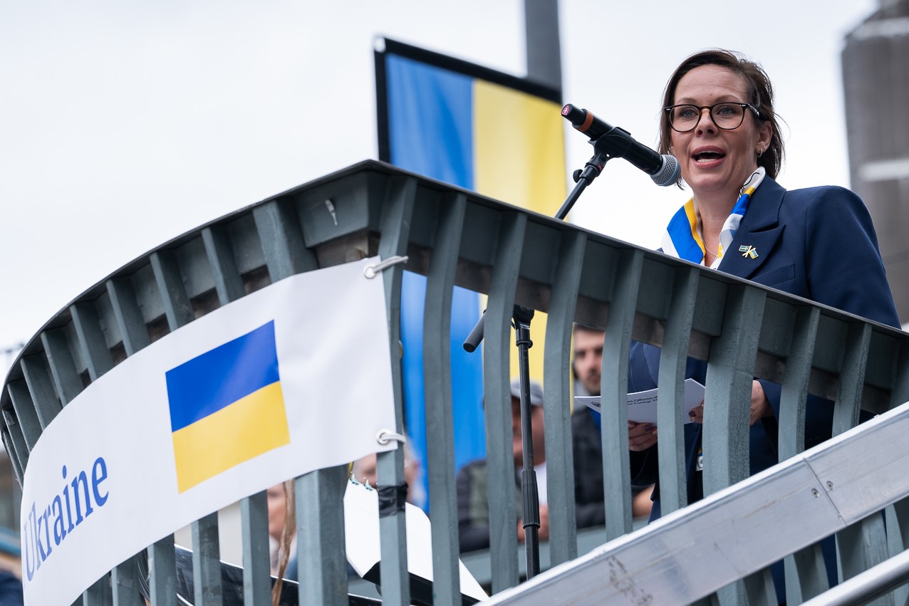 Woman speaks into microphone, Ukrainian flags in the background.
