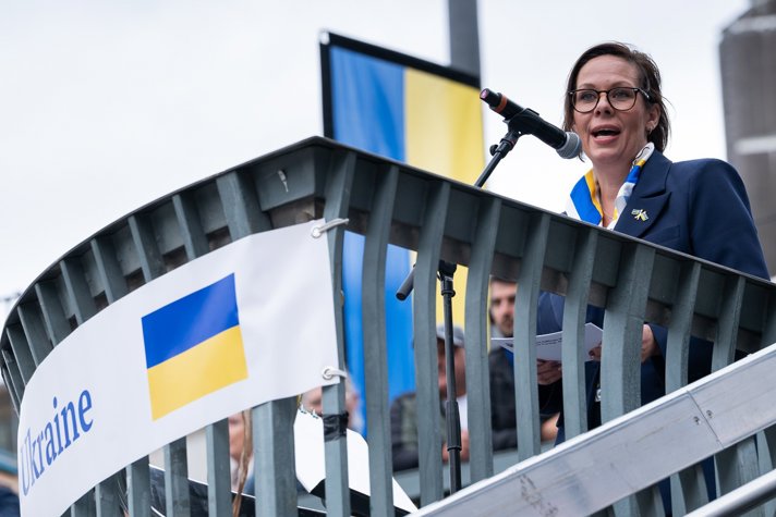 Woman speaks into microphone, Ukrainian flags in the background.