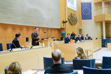 Ulf Kristersson står i talarstolen i riksdagens plenisal. I för- och bakgrunden syns åhörare.