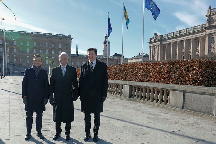 Prime Minister Ulf Kristersson, His Majesty The King and Speaker of the Riksdag Andreas Norlén standing outside the Parliament. The raised flags in the background.
