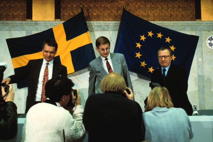 Ulf Dinkelspiel, Carl Bildt and Jaques Delors are standing in front of a table and some photographers. In the background there is a Swedish flag and a EU flag.