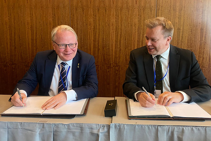 Peter Hultqvist and Antti Kaikkonen sitting next to each other and write their names  on some paper. 