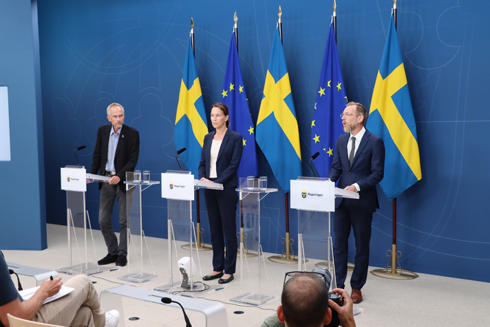 Three persons in a press conference. There are Swedish and EU flags behind them.